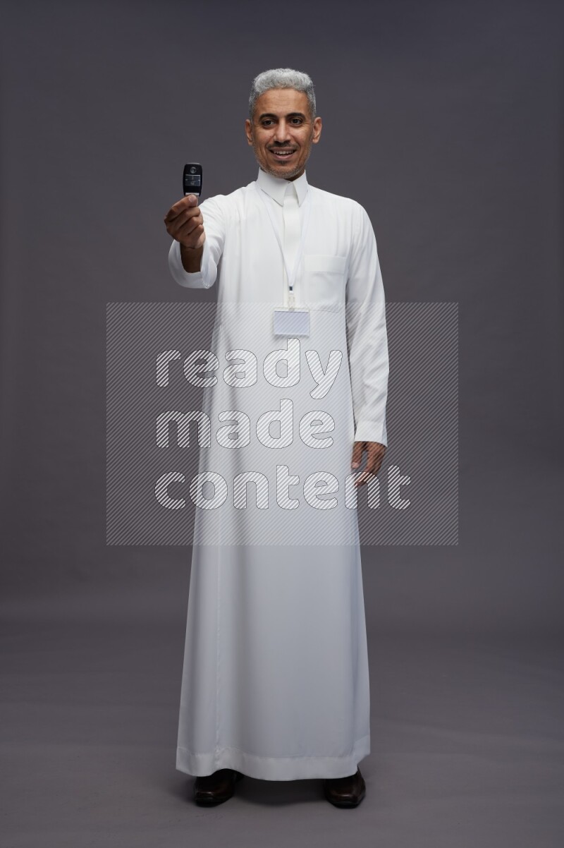 Saudi man wearing thob with neck strap employee badge standing holding key car on gray background