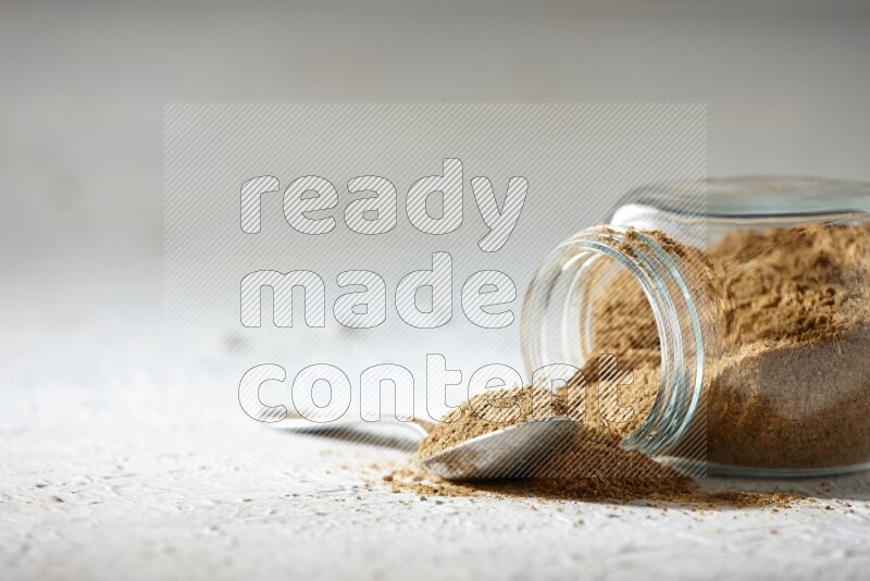 A flipped glass spice jar and a metal spoon full of cumin powder and powder spilled out on textured white flooring
