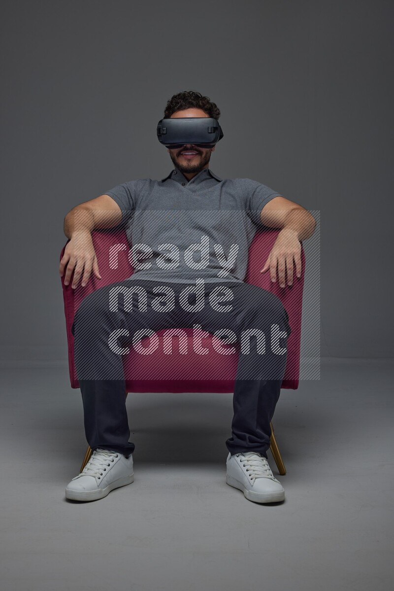 A man wearing casual and wearing VR while sitting on a burgundy chair eye level on a gray background