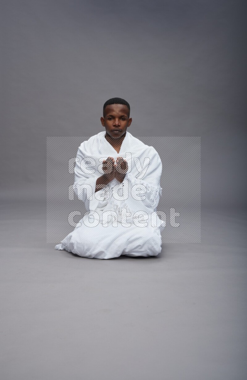 A man wearing Ehram sitting on floor dua'a on gray background