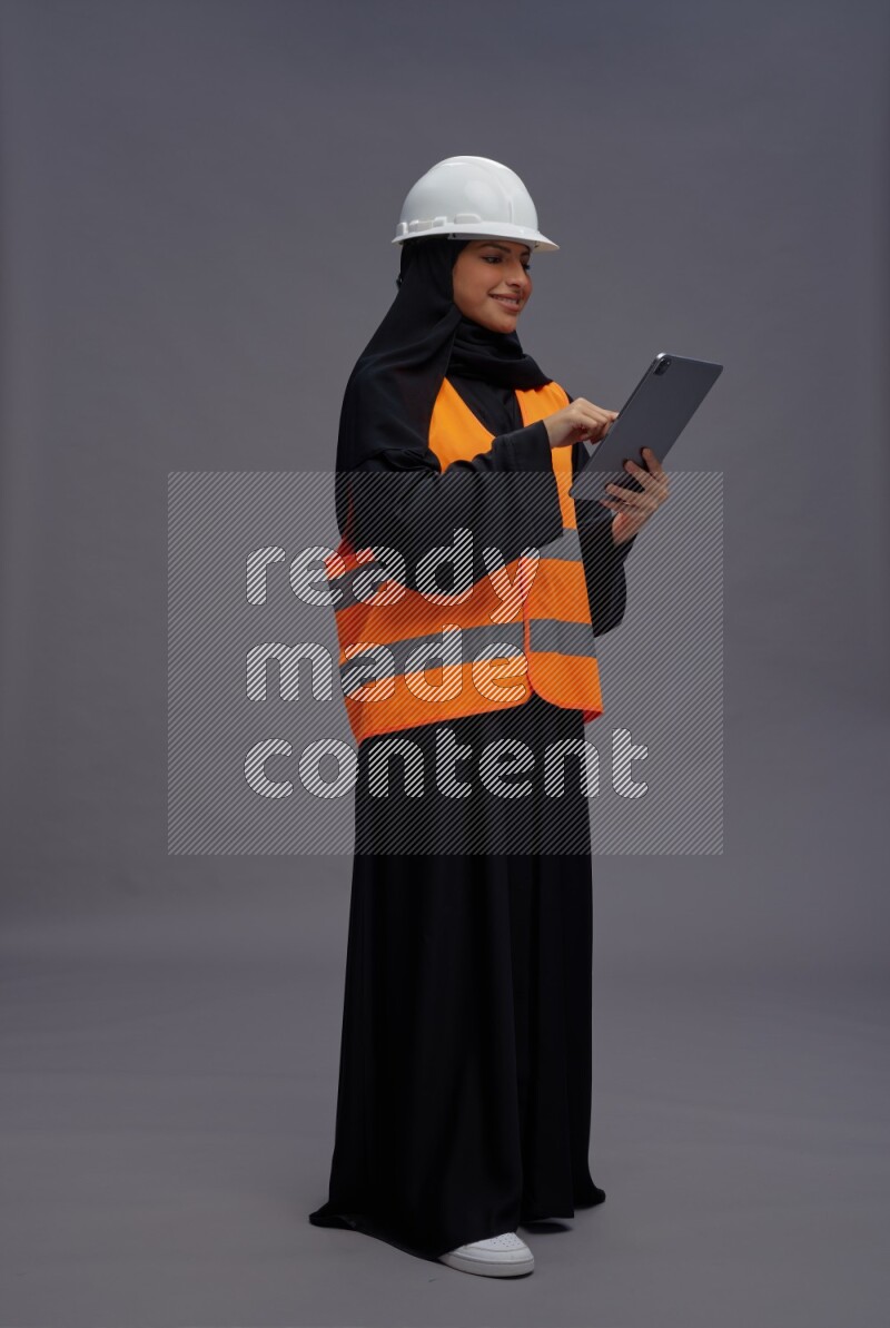 Saudi woman wearing Abaya with engineer vest standing working on tablet on gray background