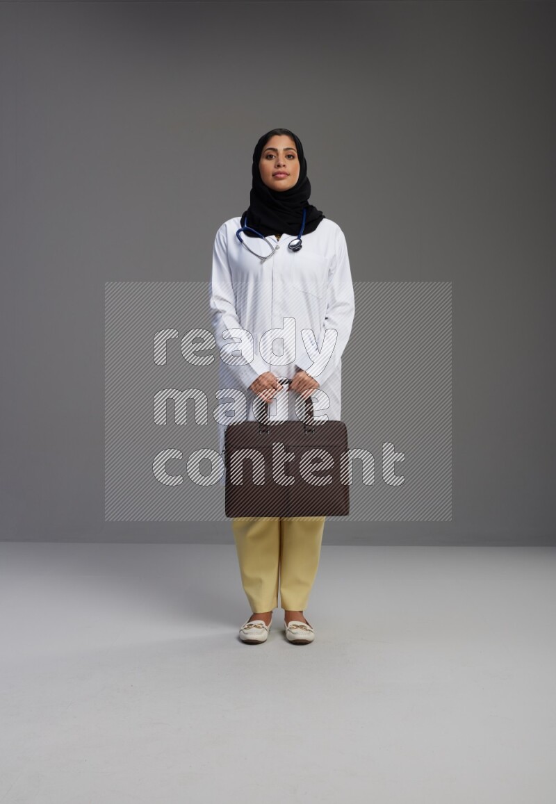Saudi woman wearing lab coat with stethoscope standing holding bag on Gray background