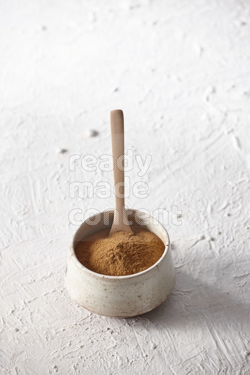 Ceramic beige bowl full of cinnamon powder with a wooden spoon on a textured white background