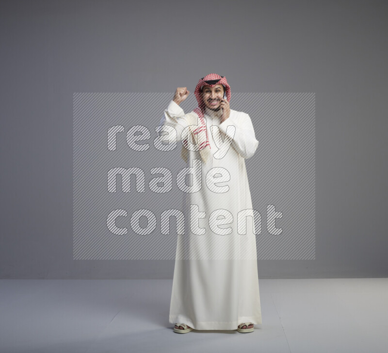 A Saudi man standing wearing thob and red shomag talking on phone on gray background