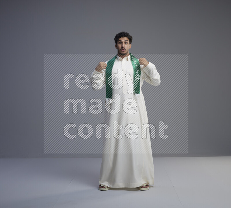 A Saudi man standing wearing thob and Saudi flag scarf on gray background