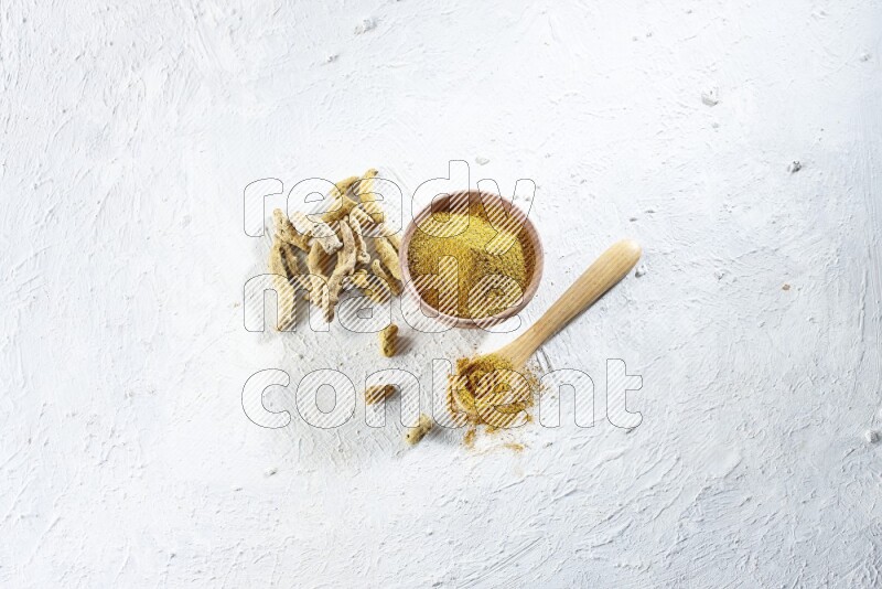 A wooden bowl and wooden spoon full of turmeric powder with dried turmeric fingers beside it on textured white flooring