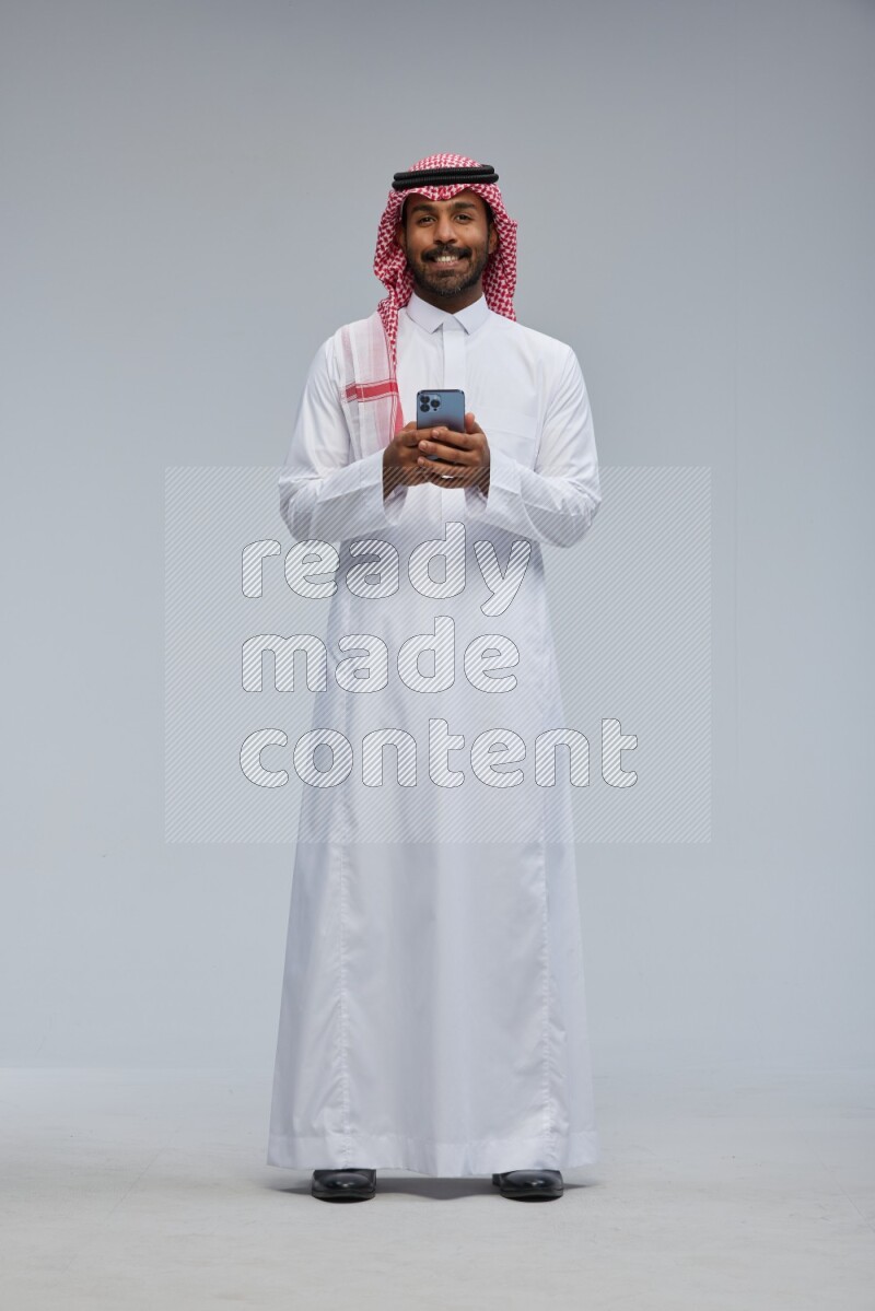 Saudi man Wearing Thob and shomag standing texting on phone on Gray background