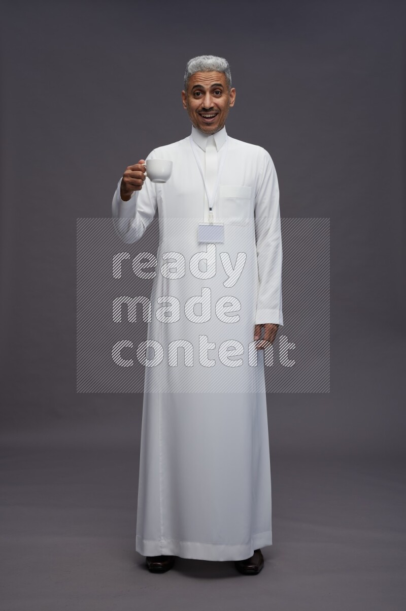 Saudi man wearing thob with neck strap employee badge standing holding mug on gray background