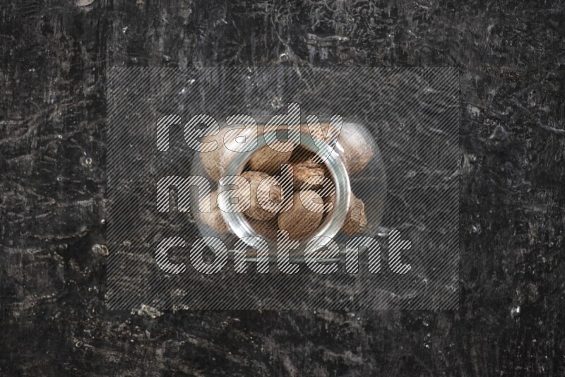 A glass spice jar full of whole nutmeg seeds on a textured black flooring