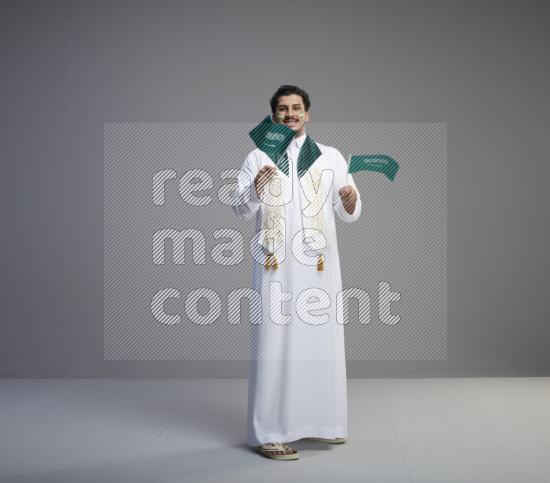 A Saudi man standing wearing thob and saudi flag scarf with face painting holding small Saudi flag on gray background