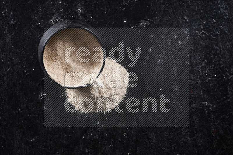 A black pottery bowl full of onion powder with fallen powder from it on black background