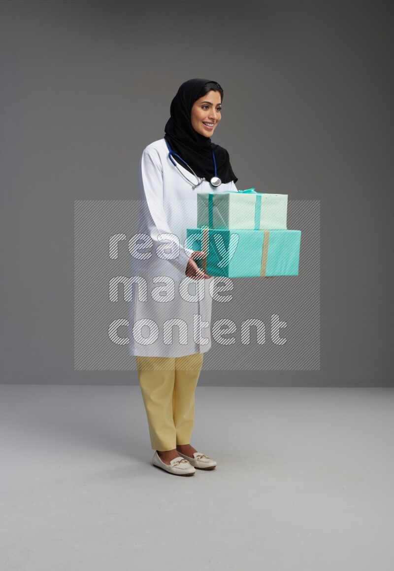 Saudi woman wearing lab coat with stethoscope standing holding gift box on Gray background