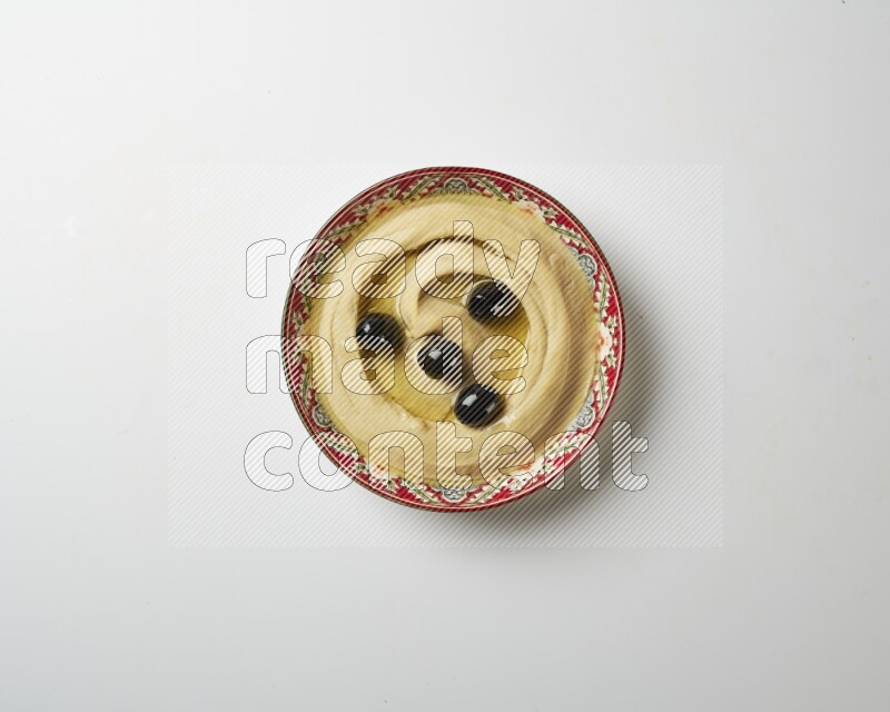 Hummus in a red plate with patterns garnished with black olives on a white background