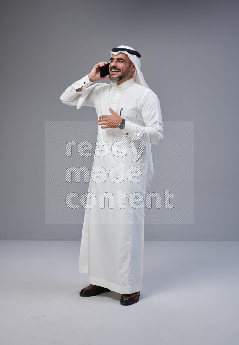 Saudi man Wearing Thob and white Shomag standing talking on phone on Gray background