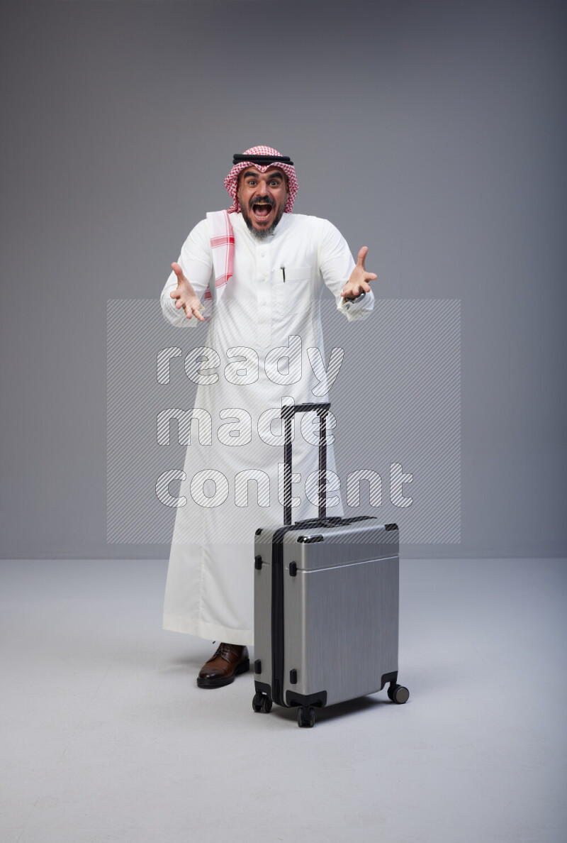 Saudi man wearing Thob and red Shomag standing holding Travel bag on Gray background