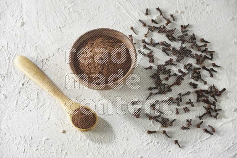 A wooden bowl and wooden spoon full of cloves powder with cloves spread on textured white flooring