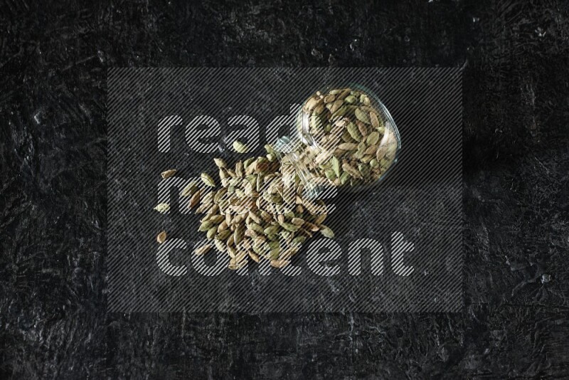 A flipped glass spice jar full of cardamom seeds on textured black flooring
