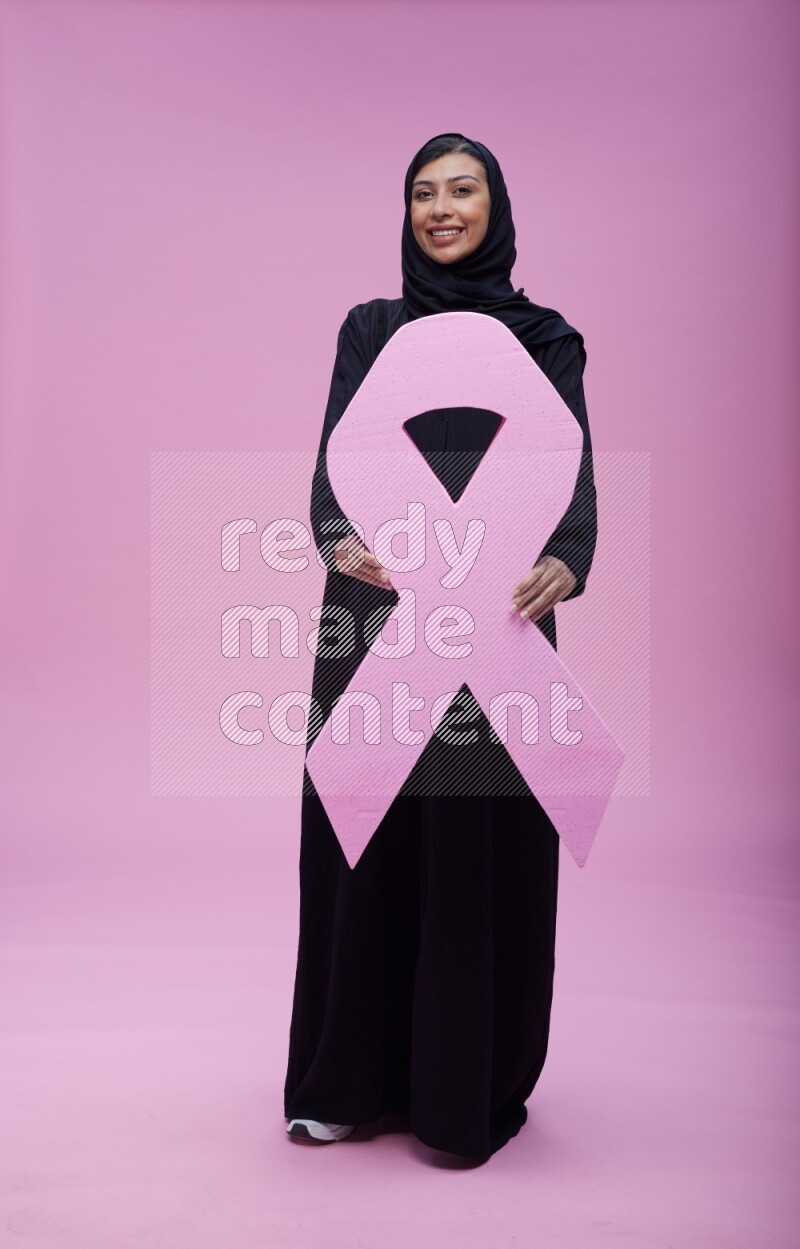 Saudi woman wearing pink ribbon on abaya standing holding awareness ribbon on plain pink background