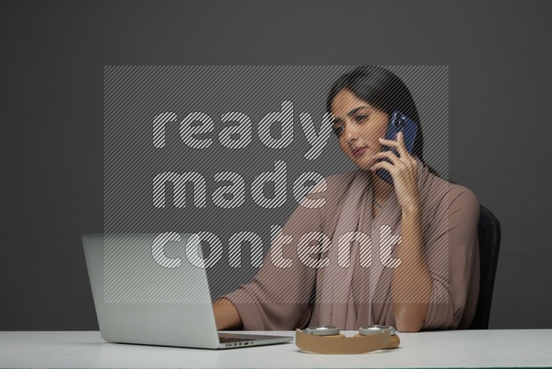 A Saudi woman Sitting on her desk Calling  on a Gray Background wearing Brown Abaya