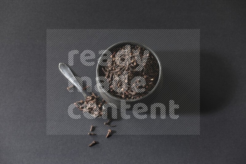A black pottery bowl full of cloves and a metal spoon next to it on a black flooring