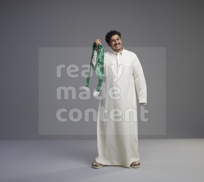 A Saudi man standing wearing thob holding Saudi flag scarf on gray background