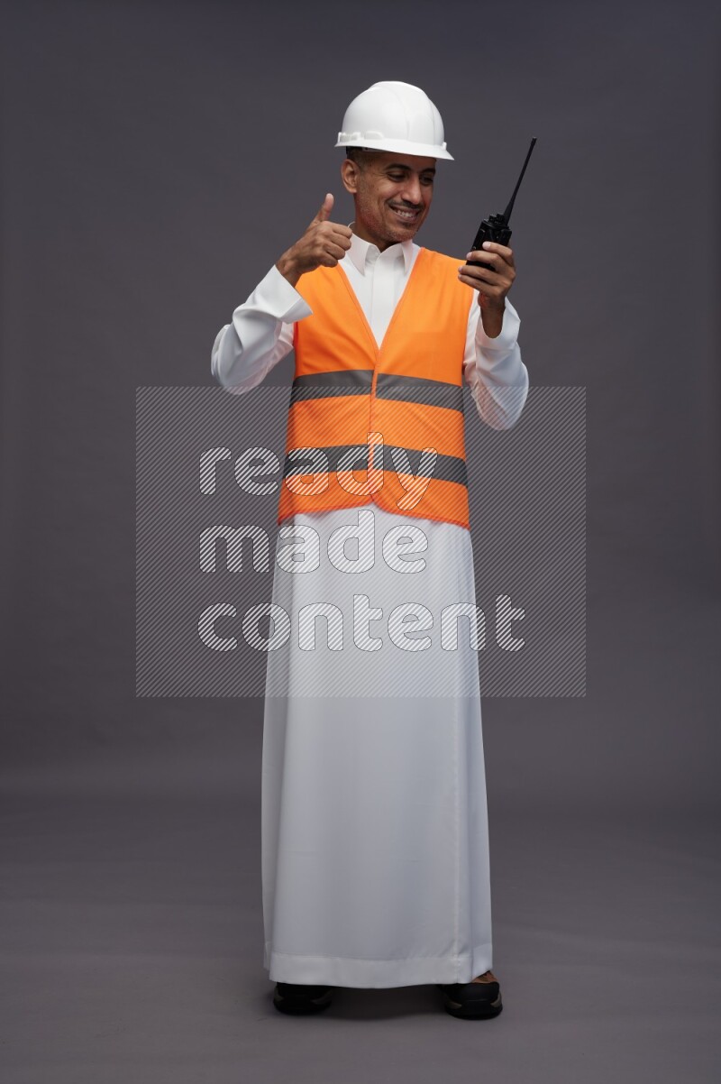 Saudi man wearing thob with engineer vest standing holding walkie-talkie on gray background