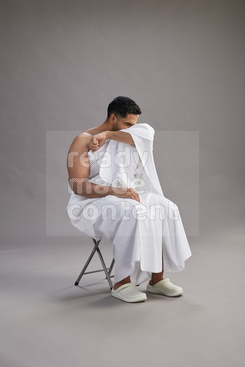 A man wearing Ehram sitting on chair showing how to sneeze on gray background