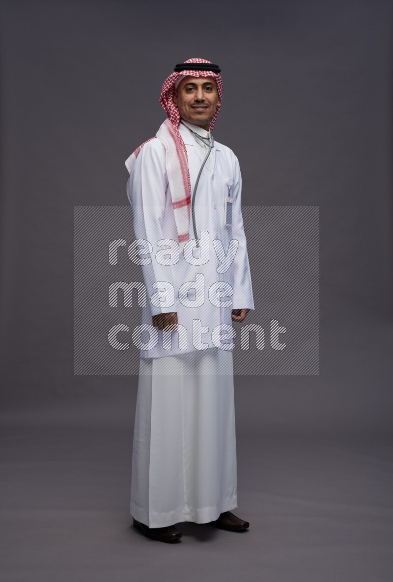 Saudi man wearing thob with lab coat and shomag with pocket employee badge with stethoscope standing interacting with the camera on gray background