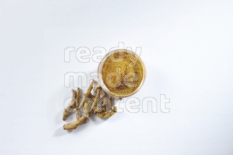 وعاء زجاجي ممتلئ ببودرة الكركم وأصابع الكركم المجففة تحته على خلفية بيضاء