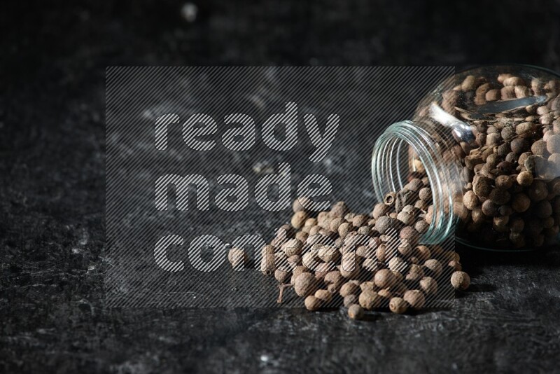 A flipped glass spice jar full of allspice whole balls and the balls spilled out of it on a textured black flooring