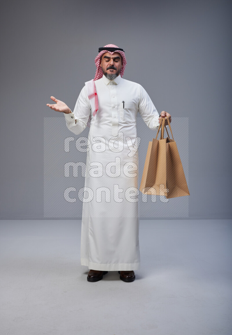 Saudi man Wearing Thob and red Shomag standing holding shopping bag on Gray background