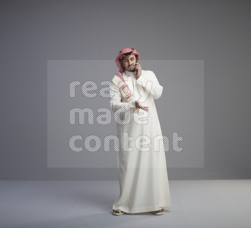 A Saudi man standing wearing thob and red shomag talking on phone on gray background