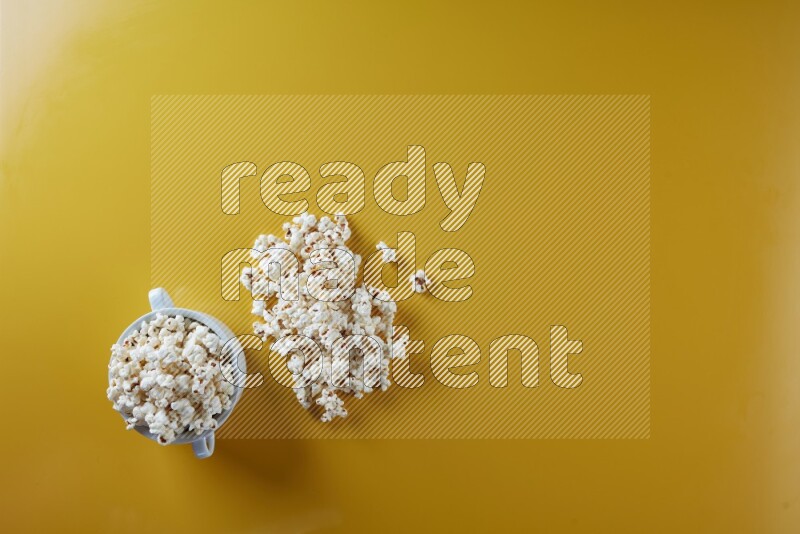 A white ceramic bowl full of popcorn with popcorn beside it on a yellow background in a top view shot