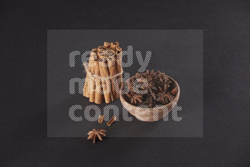 A stacked and bounded cinnamon sticks and a wooden bowl full of star anise on a black background