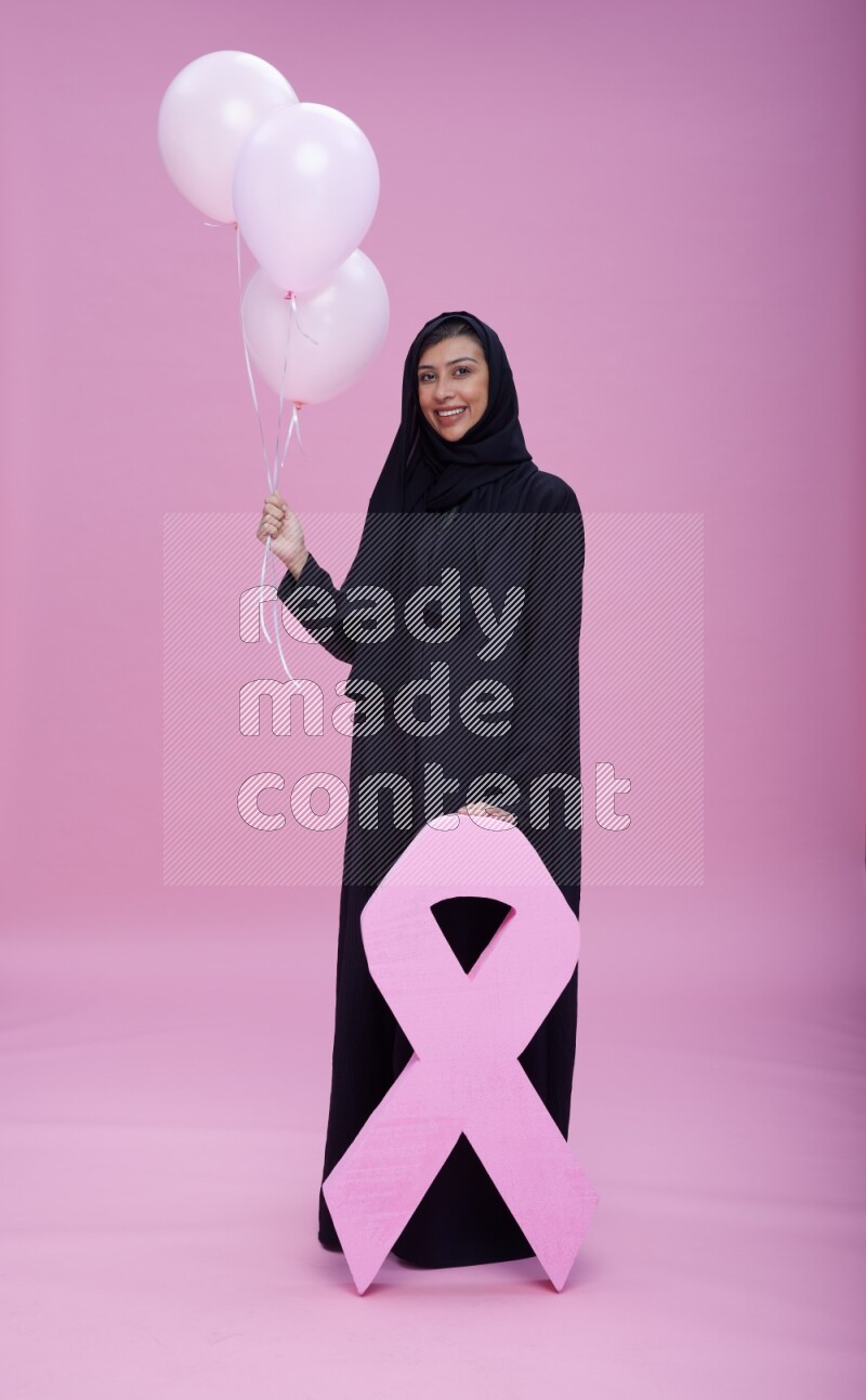 Saudi woman wearing pink ribbon on Abaya standing holding balloons and awareness sign on pink background