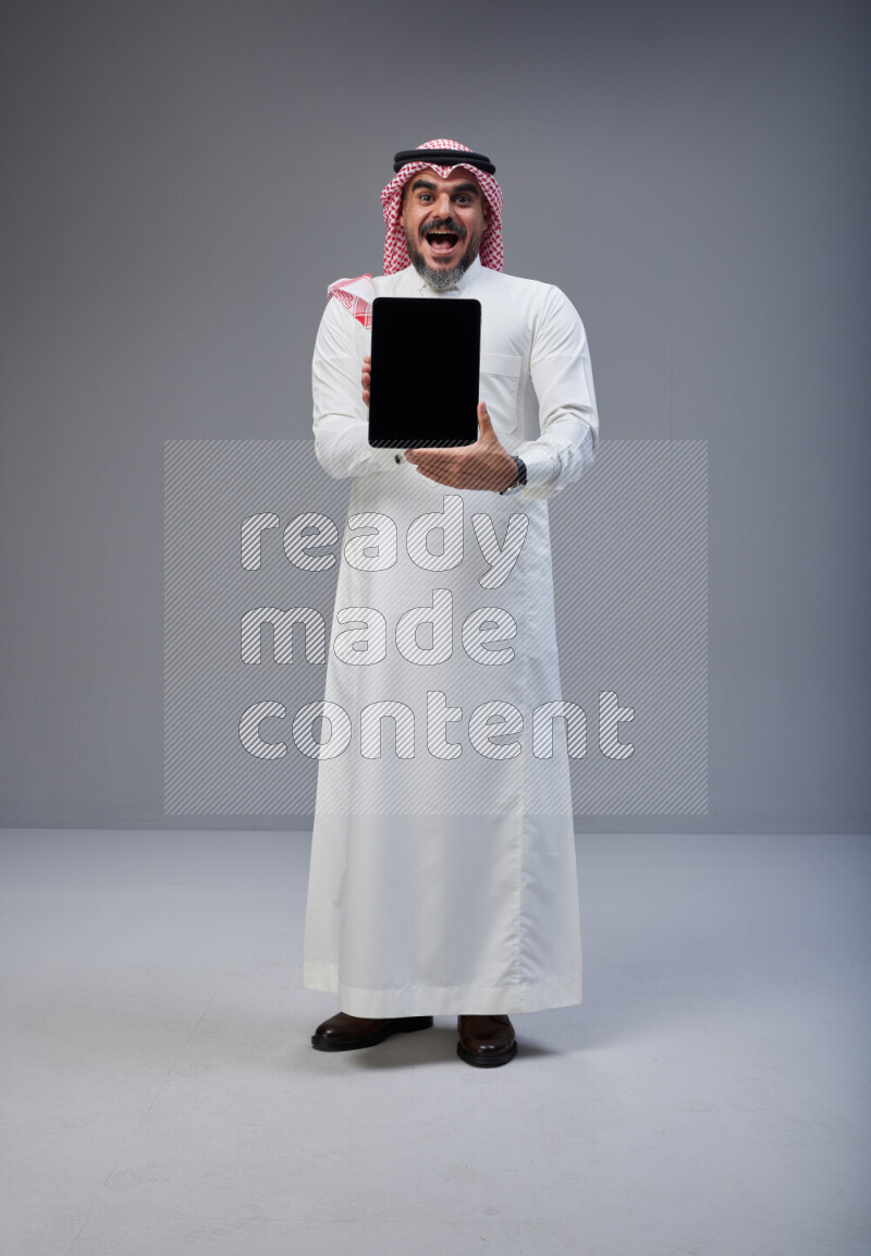 Saudi man Wearing Thob and red Shomag standing showing tablet to camera on Gray background