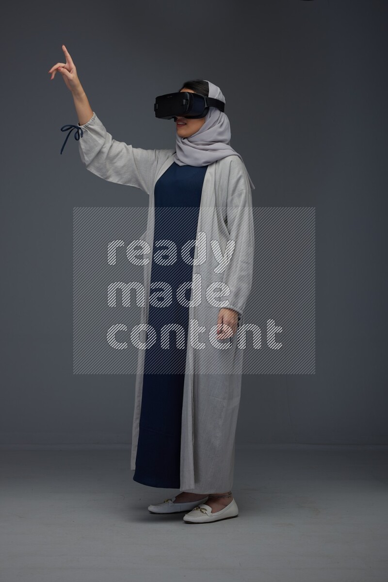 A Saudi woman wearing a light gray Abaya and head scarf standing and wearing VR eye level on a grey background