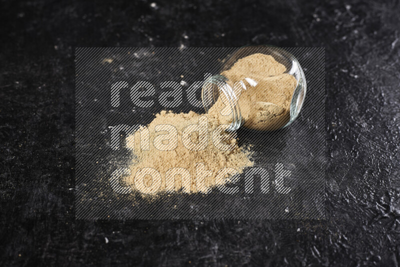 A glass jar full of ground ginger powder flipped with some spilling powder on black background