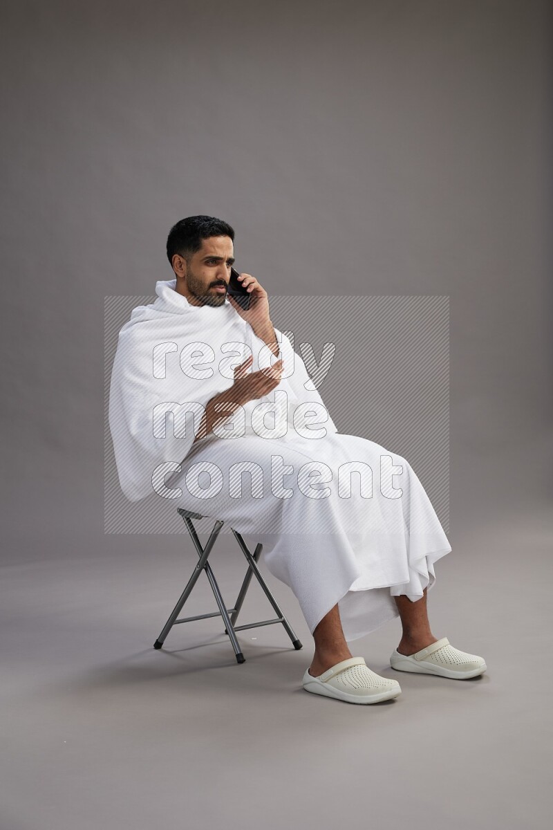 A man wearing Ehram sitting on chair talking on the phone on gray background