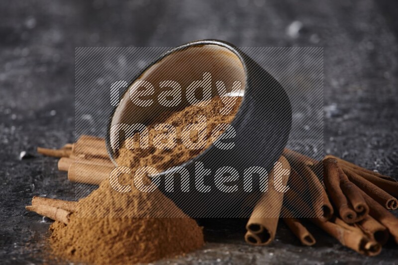 Black pottery bowl over filled with cinnamon powder and cinnamon sticks around the bowl on a textured black background