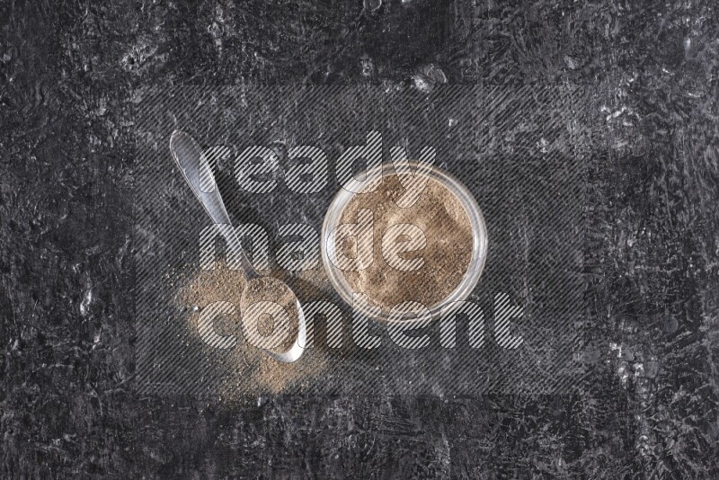 A glass jar full of black pepper powder and a metal spoon full of powder on a textured black flooring