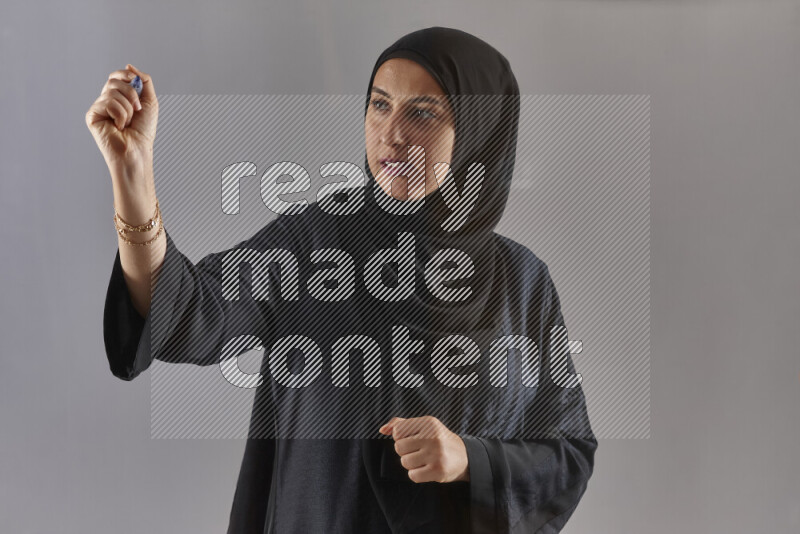 Woman in black abaya and hijab holding a marker pen to write on a transparent board with different reactions and expressions on grey background