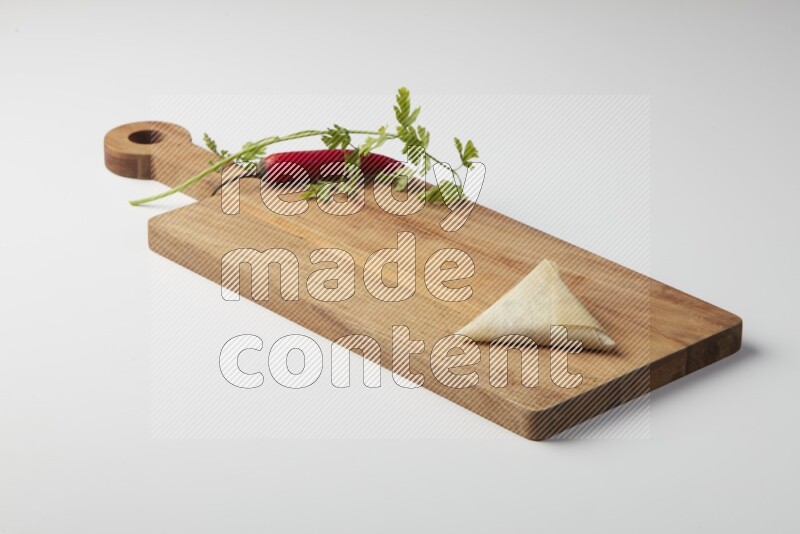 One uncooked samosa with a single red pepper and parsley on a wooden cutter on a white background