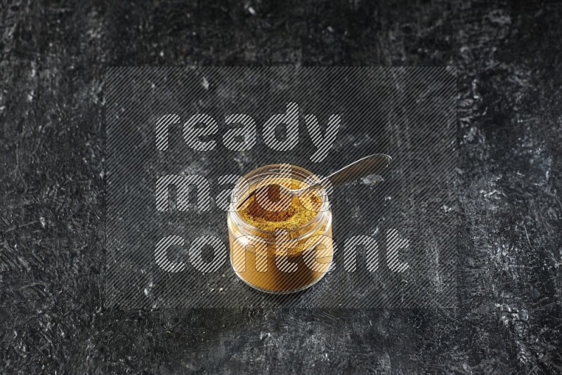 A glass jar and a metal spoon full of turmeric powder on a textured black flooring