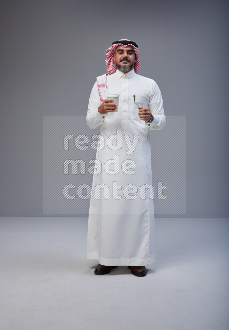Saudi man Wearing Thob and red Shomag standing holding paper cup on Gray background