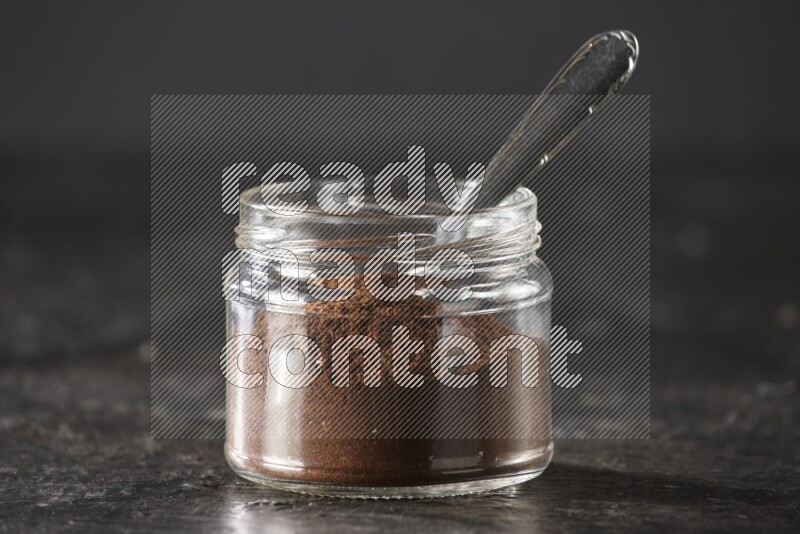 A glass jar full of cloves powder with a metal spoon on a textured black flooring