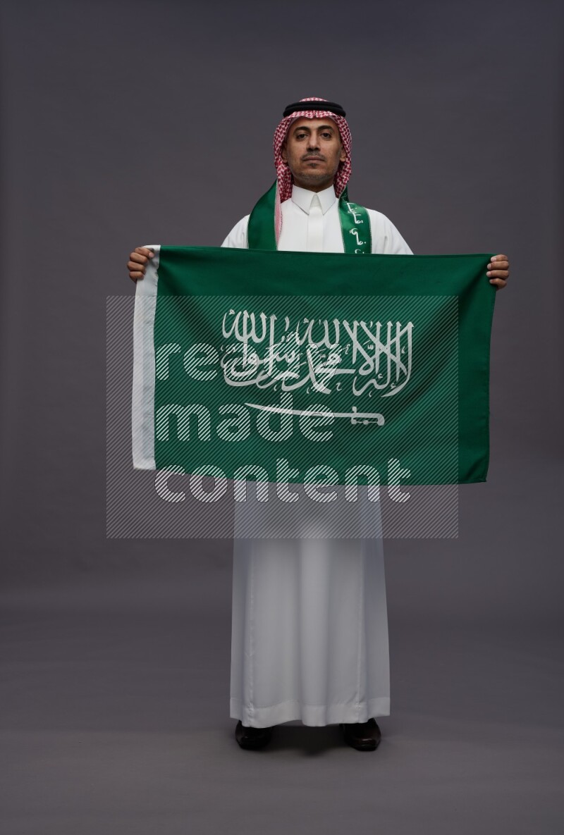 Saudi man wearing thob and shomag standing holding Saudi flag on gray background