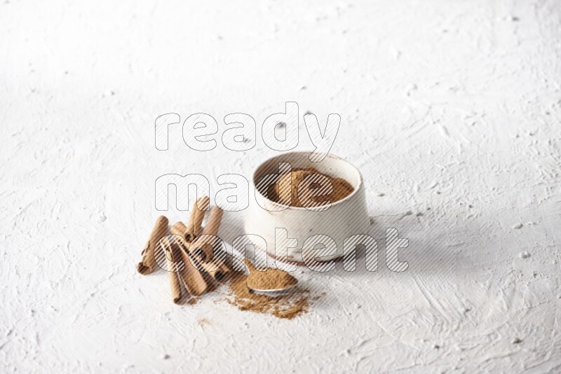 Ceramic beige bowl full of cinnamon powder and a metal spoon with cinnamon sticks next of it on a textured white background