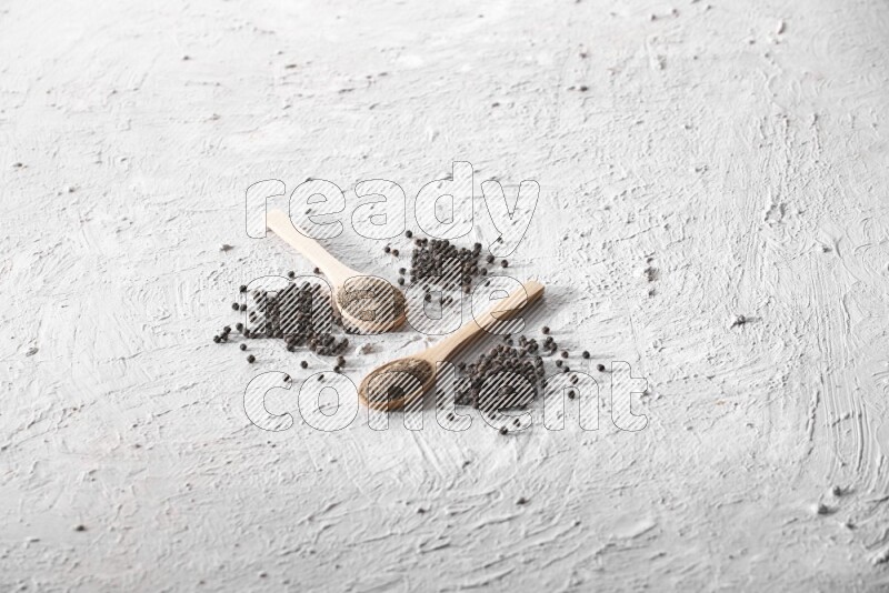 2 wooden spoons full of black pepper powder and black pepper beads spread on textured white background