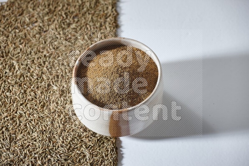 وعاء بيج ممتلئ ببودرة الكمون مع حبوب الكمون تحت الوعاء على خلفية بيضاء